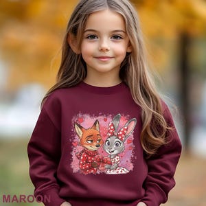 May include: A maroon sweatshirt featuring a cartoon image of a fox and a rabbit holding a heart-shaped gift. The fox is wearing a red sweater, and the rabbit has a red bow. The word "MAROON" is printed at the bottom of the sweatshirt.