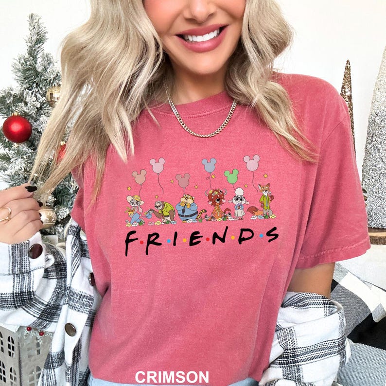 May include: Crimson-colored t-shirt featuring a cartoon design of various animal characters holding Mickey Mouse-shaped balloons. The word "FRIENDS" is printed in black below the characters. The shirt is a short-sleeved crew neck.