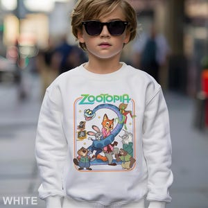 May include: White sweatshirt featuring the word "Zootopia" in green and a colorful graphic of characters from the animated movie. The design includes a fox, rabbit, and other animals. The sweatshirt is worn by a person wearing sunglasses.