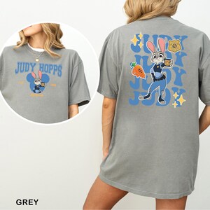 Op de afbeelding: Een grijs t-shirt met een afbeelding van Judy Hopps op de achterkant en een kleinere versie op de voorkant. Het ontwerp op de achterkant bevat het personage, een wortel, een politiebadge en de tekst Judy herhaald in het blauw. De voorkant toont Judy Hopps in een blauw muizenoorsilhouet met de tekst Judy Hopps.