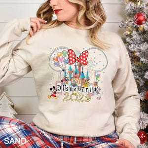 Puede incluir: Una sudadera de color arena con un dise&ntilde;o de castillo de Disney colorido con Mickey y Minnie Mouse, el Pato Donald y otros personajes. El dise&ntilde;o incluye el texto "Disney Trip 2020" en letras doradas, con un lazo rojo de lunares.