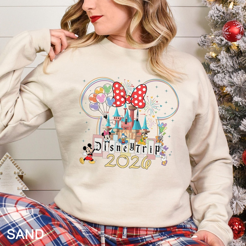 Puede incluir: Una sudadera de color arena con un dise&ntilde;o de castillo de Disney colorido con Mickey y Minnie Mouse, el Pato Donald y otros personajes. El dise&ntilde;o incluye el texto "Disney Trip 2020" en letras doradas, con un lazo rojo de lunares.