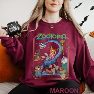 May include: Maroon sweatshirt featuring a colorful graphic from the movie Zootopia. The design includes the movie title, characters, and a large number 2. The word "MAROON" is printed at the bottom of the sweatshirt.