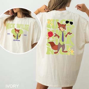 Op de afbeelding: Een ivoorkleurig t-shirt met een afbeelding van het personage Nick Wilde uit Zootopia. De achterkant toont een grote illustratie van de vos met een rood pootvormig ijsje, een zonnebril en de naam Nick herhaald in het groen op de achtergrond.