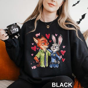 Pu&ograve; includere: Felpa nera con un'illustrazione colorata di Nick Wilde e Judy Hopps di Zootropolis, circondati da cuori rossi. La parola "BLACK" &egrave; stampata in basso. Nella foto si tiene una tazza nera con la scritta "Witches Brew".
