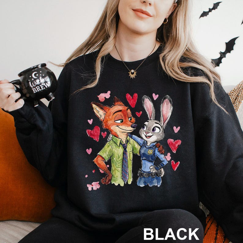 Pu&ograve; includere: Felpa nera con un'illustrazione colorata di Nick Wilde e Judy Hopps di Zootropolis, circondati da cuori rossi. La parola "BLACK" &egrave; stampata in basso. Nella foto si tiene una tazza nera con la scritta "Witches Brew".