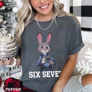 May include: Dark grey t-shirt with a cartoon image of Judy Hopps from Zootopia, in a police uniform, making a hand gesture. The text "SIX SEVE'" is printed in white below the image. The shirt is a comfortable, casual style.
