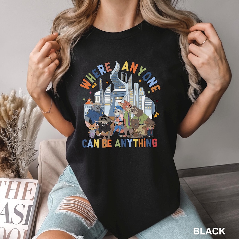 May include: Black t-shirt with a colorful graphic featuring cartoon animal characters and the text "WHERE ANYONE CAN BE ANYTHING". The design includes a cityscape and various animal figures.