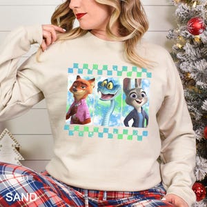 May include: Beige sweatshirt featuring three cartoon characters: a fox in a pink shirt, a blue snake, and a rabbit in a blue jacket. The design is framed with a blue and green checkered pattern. The sweatshirt is part of a casual wear collection.