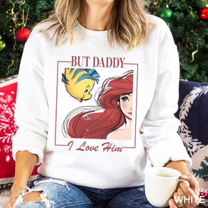 May include: White crew neck sweatshirt with a framed illustration of Ariel and Flounder from The Little Mermaid. The maroon text reads "BUT DADDY - I Love Him". The sweatshirt has long sleeves.