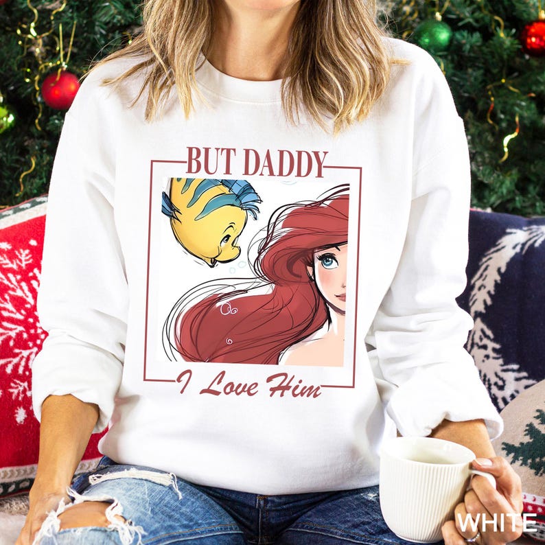 May include: White crew neck sweatshirt with a framed illustration of Ariel and Flounder from The Little Mermaid. The maroon text reads "BUT DADDY - I Love Him". The sweatshirt has long sleeves.