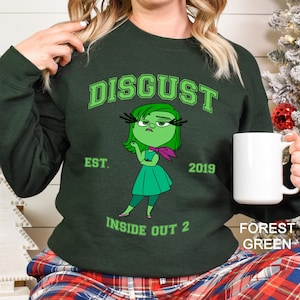 May include: Forest green sweatshirt featuring the word "DISGUST" in green block letters above a cartoon character from the movie "Inside Out 2". The character is green with a pink scarf and a matching green dress. The text "EST. 2019" and "INSIDE OUT 2" are also printed on the sweatshirt.