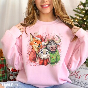 May include: Light pink sweatshirt featuring a graphic of a fox, bunny, and bull in Christmas attire. The fox wears a Santa hat and red coat, the bunny has red mittens, and the bull wears a green sweater. The sweatshirt has a crew neck.