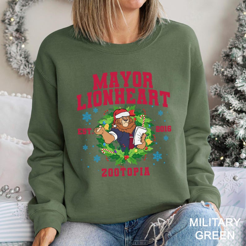 Pu&ograve; includere: Felpa verde militare con la scritta rossa "MAYOR LIONHEART" e un leone dei cartoni animati con un cappello da Babbo Natale. Il design include una ghirlanda, fiocchi di neve, la parola "ZOOTOPIA" e l'anno "2016".