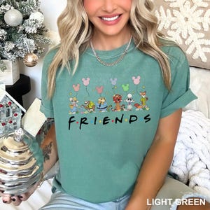 May include: Light green t-shirt with the word "FRIENDS" in black letters. The shirt features cartoon characters holding balloons. The balloons are in the shape of Mickey Mouse ears. The shirt is a light green color.