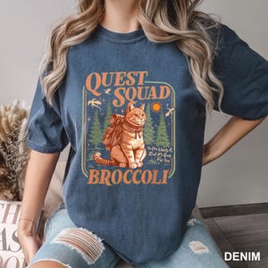 Pode incluir: Camiseta azul denim com um gr&aacute;fico de um gato com uma mochila, o texto "QUEST SQUAD BROCCOLI" e a frase "No One Wants It, But It's Good For You". O gato &eacute; laranja e branco.