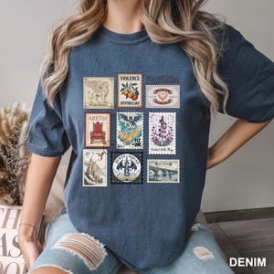 May include: A denim blue t-shirt featuring a collection of postage stamp designs. The stamps showcase various illustrations, including a griffin, a dragon, and a bridge. Text on the stamps includes "Violence," "Apothecary," and "Quest Squad."