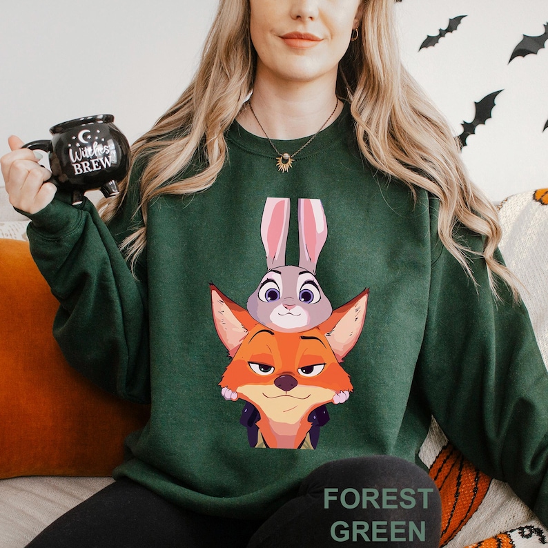 May include: Forest green sweatshirt featuring a cartoon fox and bunny design. The fox is orange and the bunny is gray. The text "FOREST GREEN" is printed at the bottom of the sweatshirt. The sweatshirt is a long-sleeved crew neck.