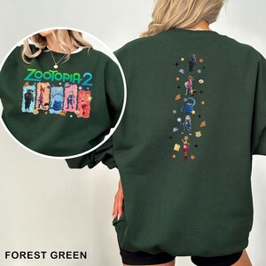K&ouml;nnte beinhalten: Forstgr&uuml;nes Sweatshirt mit dem Text "ZOOTOPIA 2" und Charaktergrafiken auf der Vorderseite. Die R&uuml;ckseite hat ein vertikales Design mit Charakterillustrationen und Sternen. Das Sweatshirt hat einen Rundhalsausschnitt.