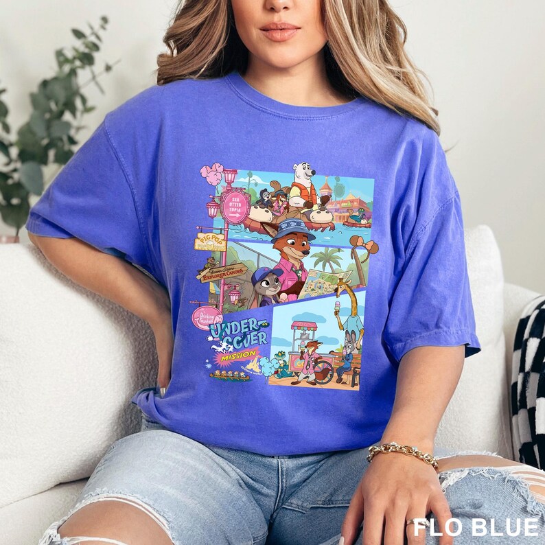 May include: A periwinkle blue short-sleeve t-shirt featuring a colorful graphic print of various animated animal characters in a theme park setting. The design includes multiple scenes with characters, palm trees, and the text Undercover Mission.