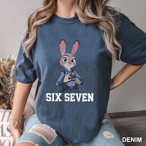 May include: A denim blue t-shirt with a cartoon image of Judy Hopps from Zootopia. The character is in a police uniform and making a hand gesture. The text "SIX SEVEN" is printed in white below the image.