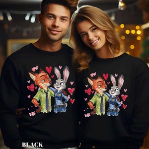 Pu&ograve; includere: Felpe nere con un cartone animato di una volpe e un coniglio che si abbracciano, circondati da cuori rossi. La volpe indossa una camicia verde e il coniglio un'uniforme blu. La parola "BLACK" &egrave; stampata in basso.