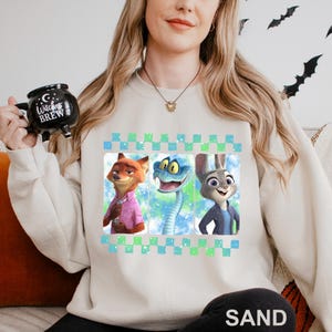 May include: Sand-colored sweatshirt featuring a graphic of three cartoon characters: a fox in a pink shirt, a snake, and a rabbit in a blue jacket. The design is framed with a blue and green checkered pattern. The word "SAND" is printed at the bottom.