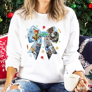 May include: White sweatshirt featuring a large blue bow design with characters from the movie "Zootopia". The bow is filled with various characters and the word "Zootopia" in green. The sweatshirt is a long-sleeved crew neck.
