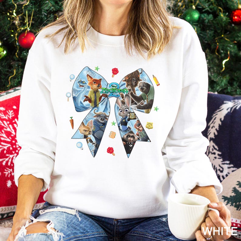 May include: White sweatshirt featuring a large blue bow design with characters from the movie "Zootopia". The bow is filled with various characters and the word "Zootopia" in green. The sweatshirt is a long-sleeved crew neck.