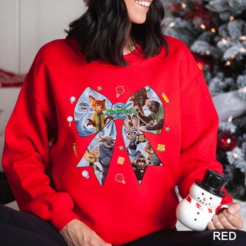 May include: A red sweatshirt featuring a large bow design with characters from the movie "Zootopia." The bow includes images of Nick Wilde, Judy Hopps, and other characters. The word "Zootopia" is printed on the bow.