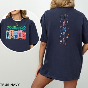 Op de afbeelding: Marineblauw T-shirt met de tekst "ZOOTOPIA 2" en karakterillustraties op de voorkant. De achterkant van het shirt heeft verticale illustraties van karakters en sterren.