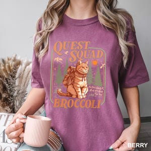 Pode incluir: Camiseta cor de baga com um gr&aacute;fico de um gato com uma mochila, as palavras "Quest Squad" e "Broccoli". O design inclui &aacute;rvores, um sol e o texto "No One Wants It, But It's Good For You".