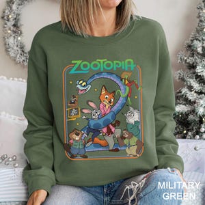 May include: Military green sweatshirt featuring a colorful graphic from the animated film "Zootopia." The design includes the film's title and various characters, such as a fox, rabbit, and sloth. The sweatshirt is a crew neck style.