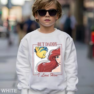 May include: White sweatshirt with a graphic of Ariel and Flounder from The Little Mermaid, and the text "BUT DADDY - I Love Him" in a red frame. The sweatshirt is worn by a child.