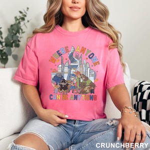May include: Pink t-shirt with a colorful graphic featuring cartoon characters and the text "WHERE ANYONE CAN BE ANYTHING". The design includes a cityscape and various animal figures. The shirt is a solid pink color.