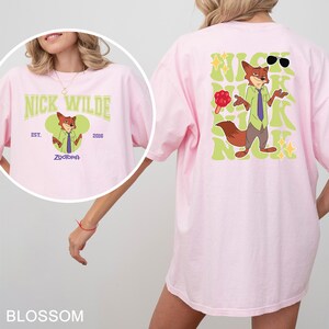 Op de afbeelding: Een lichtroze t-shirt met een afbeelding van het personage Nick Wilde uit Zootropolis. De voorkant toont de tekst Nick Wilde, Est. 2016 en Zootopia. De achterkant toont een grote illustratie van Nick Wilde met de herhaalde tekst Nick.