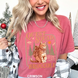 Pode incluir: Camiseta carmesim com um gr&aacute;fico com um gato usando uma mochila, as palavras "Quest Squad" e "Broccoli". O design inclui &aacute;rvores, um sol e o texto "No One Wants It, But It's Good For You".