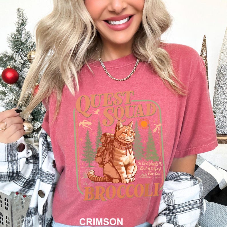 Pode incluir: Camiseta carmesim com um gr&aacute;fico com um gato usando uma mochila, as palavras "Quest Squad" e "Broccoli". O design inclui &aacute;rvores, um sol e o texto "No One Wants It, But It's Good For You".