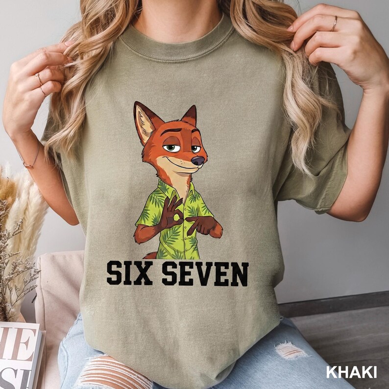 May include: Khaki t-shirt with a cartoon fox in a green Hawaiian shirt, gesturing the 'okay' sign. The words 'SIX SEVEN' are printed in bold black font beneath the character. The shirt is a casual style, suitable for everyday wear.