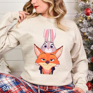 May include: A beige sweatshirt featuring a graphic of the characters Judy Hopps and Nick Wilde from the animated film Zootopia. Judy, a gray rabbit, is atop Nick, an orange fox. The word "SAND" is printed in the lower left corner.