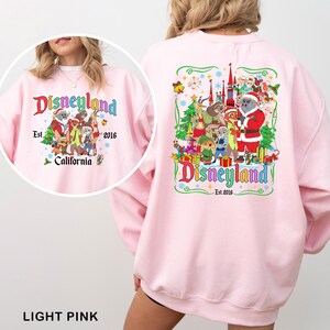May include: Light pink Disneyland sweatshirt with a colorful graphic on the front and back. The front graphic says "Disneyland Est. 2016 California" with cartoon characters. The back graphic has a similar design with the Disneyland logo.