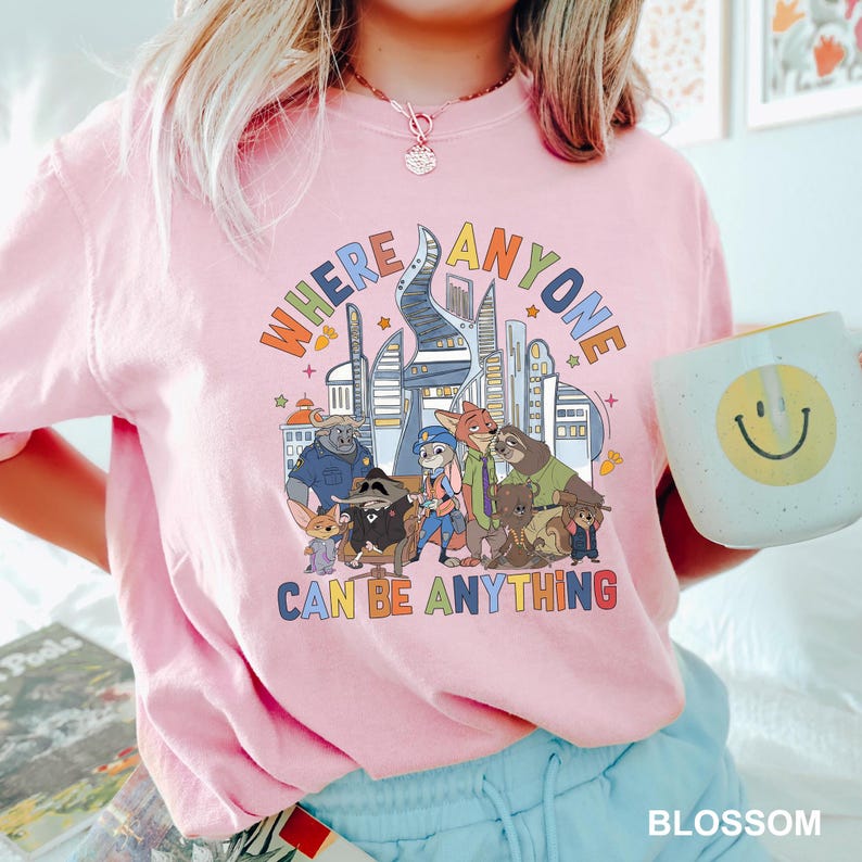May include: A light pink t-shirt with the text "WHERE ANYONE CAN BE ANYTHING" in a colorful, retro font. The graphic features cartoon characters and a cityscape. A white mug with a yellow smiley face is held in the image.