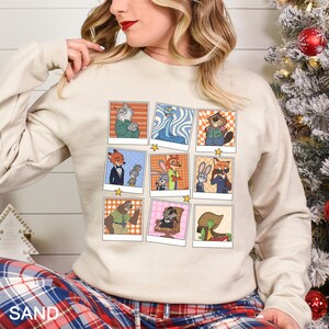 May include: Sand-colored sweatshirt featuring a grid of cartoon character portraits. Each portrait is framed with a white border and set against a light orange and white checkered background. The characters are from the animated movie Zootopia.