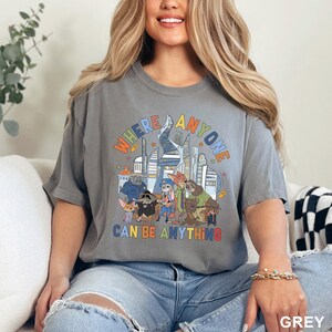 May include: A grey t-shirt with the text "WHERE ANYONE CAN BE ANYTHING" in colorful letters. The graphic features cartoon animal characters in front of a city skyline. The shirt is a unisex style.