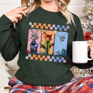 May include: Forest green sweatshirt featuring a colorful graphic of characters from the animated movie Zootopia. The design includes Judy Hopps, Nick Wilde, and a snake, framed by a checkered border. The text "FOREST GREEN" is printed at the bottom.