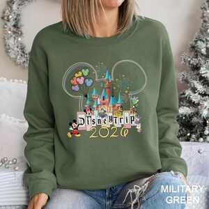 Puede incluir: Una sudadera verde oliva con un dise&ntilde;o colorido de Disney Trip 2026. El dise&ntilde;o incluye un castillo, Mickey Mouse y otros personajes de Disney. La sudadera tiene cuello redondo y mangas largas.