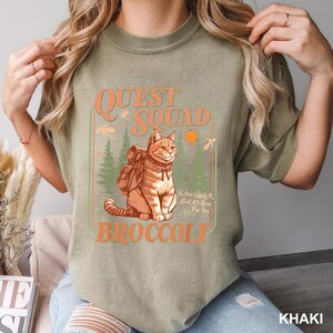 Pode incluir: Camiseta c&aacute;qui com um gr&aacute;fico que apresenta um gato com uma mochila, as palavras "QUEST SQUAD BROCCOLI" e o texto "No One Wants It, But It's Good For You". O design inclui &aacute;rvores e um sol.