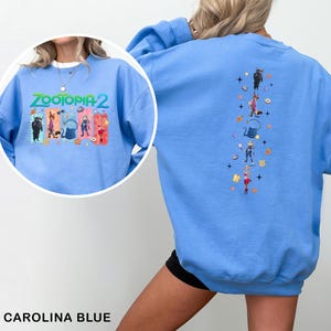 K&ouml;nnte beinhalten: Carolina-blaues Sweatshirt mit dem Text "ZOOTOPIA 2" und Cartoon-Charakteren auf der Vorderseite. Die R&uuml;ckseite hat ein vertikales Design mit Cartoon-Charakteren und Sternen. Das Sweatshirt ist aus weichem Material.
