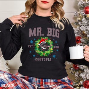 Pu&ograve; includere: Felpa nera con la scritta "MR. BIG" sopra un disegno a ghirlanda con un procione con un cappello da Babbo Natale. La ghirlanda include fiocchi di neve e la parola "ZOOTOPIA". La felpa fa parte di una collezione natalizia.