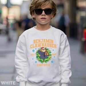 Pu&ograve; includere: Felpa bianca con la scritta "BENJAMIN CLAWHAUSER" in arancione e un'illustrazione di un personaggio di Zootopia. Il design include fiocchi di neve, l'anno 2016 e la parola "ZOOTOPIA".